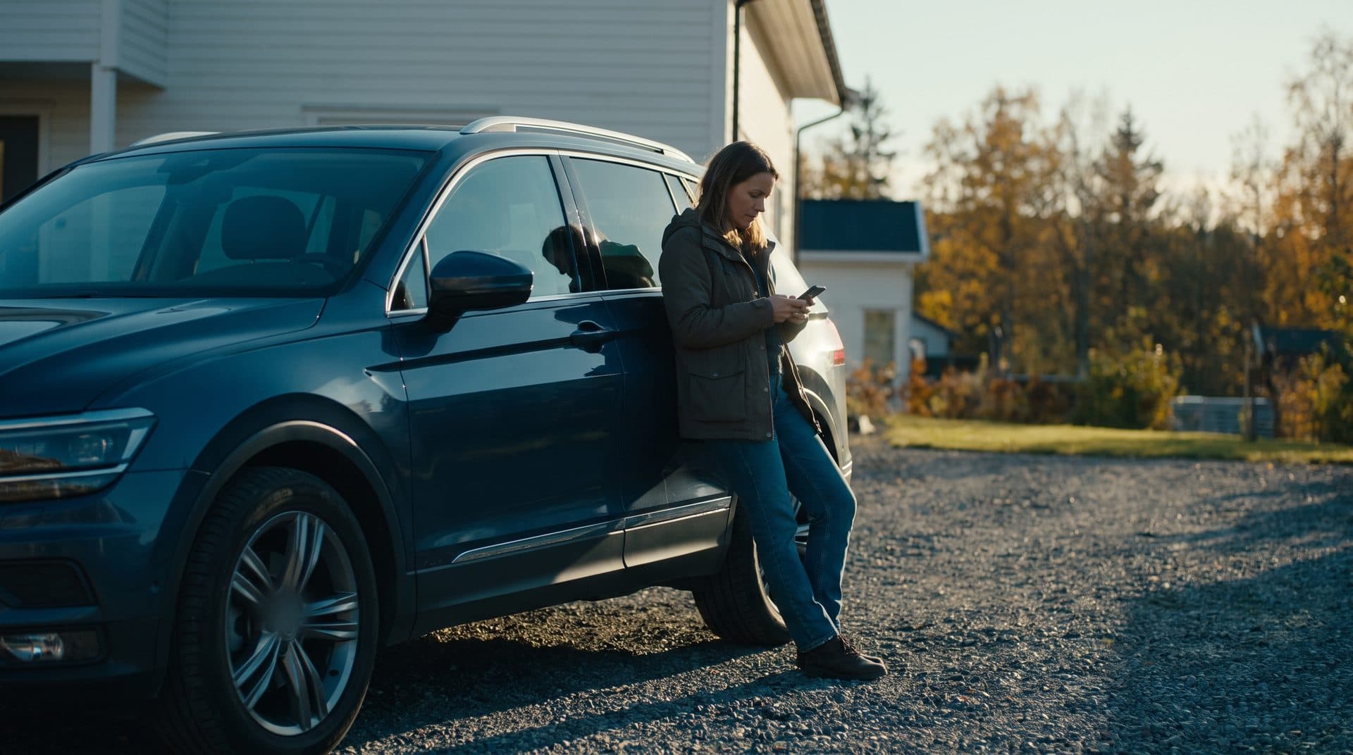 Kvinne lener seg mot den blå SUV-en sin i oppkjørselen og sjekker verdien på mobilen